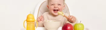 Bébé blanc et blond sur sa chaise haute en train de manger et rire.