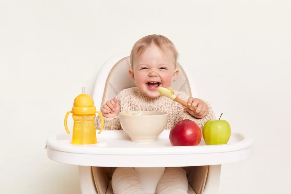 Bébé blanc et blond sur sa chaise haute en train de manger et rire.