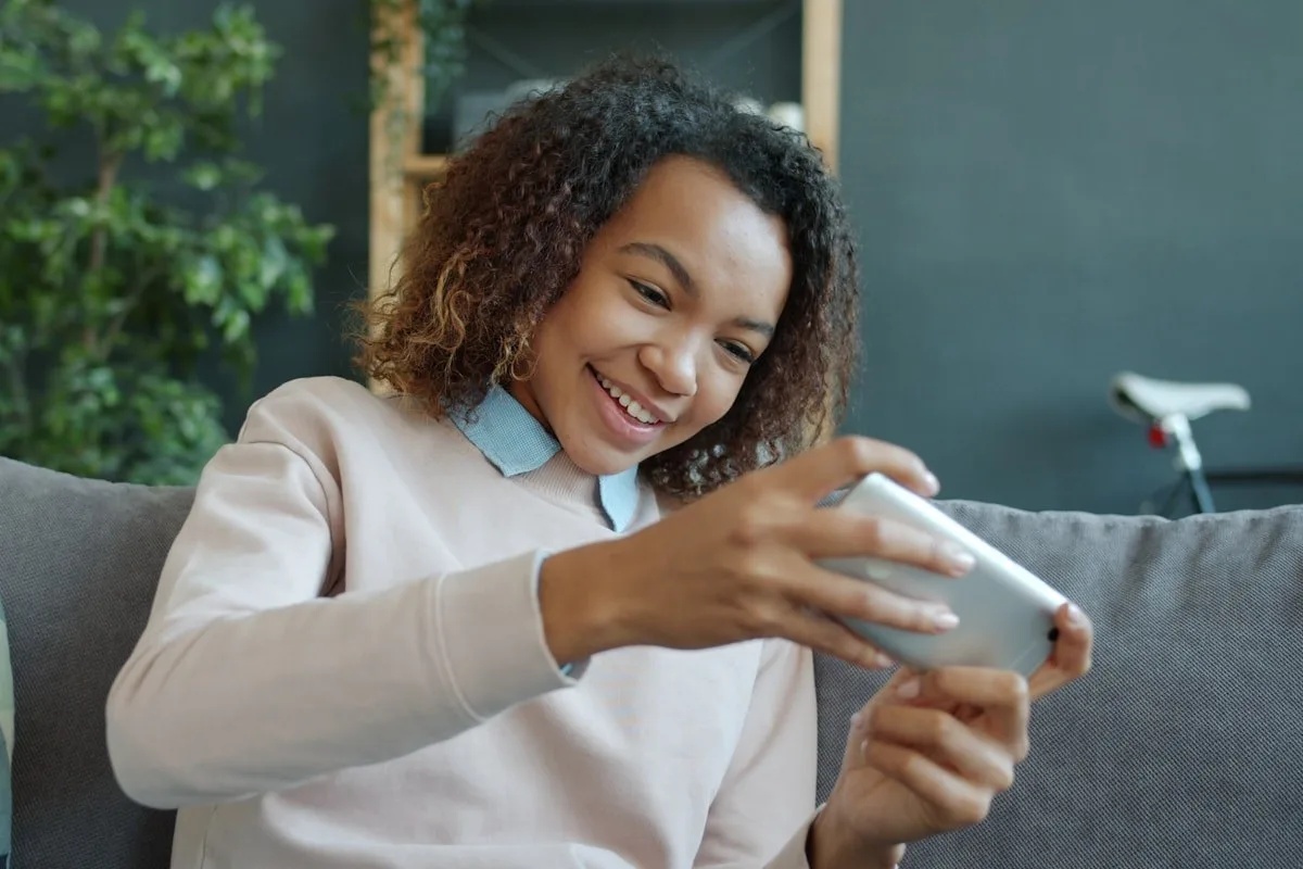 Adolescente souriante avec cheveux bouclés utilisant un smartphone assise sur un canapé gris