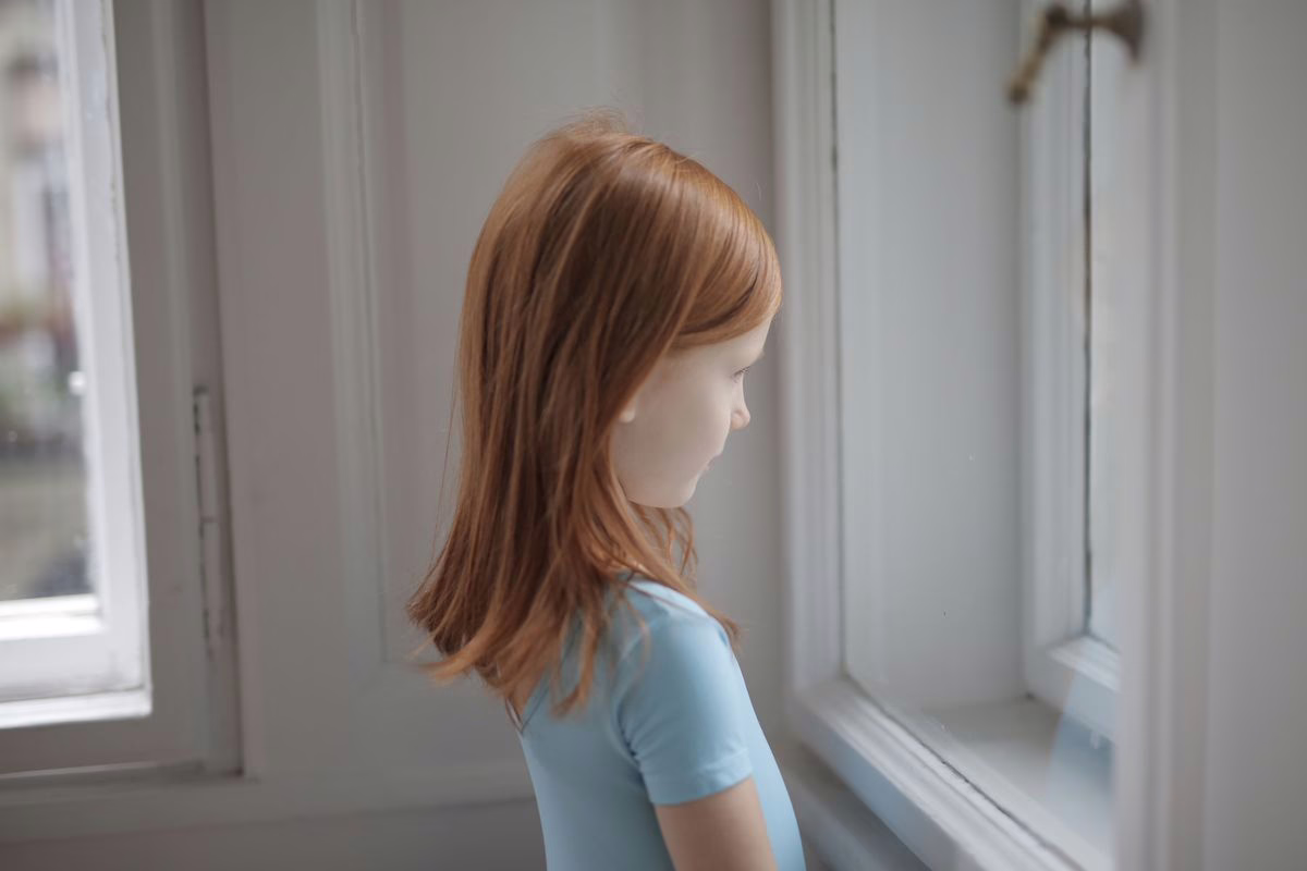 Enfant regardant par la fenêtre du salon, illustration de la question de l'enfant seul à la maison