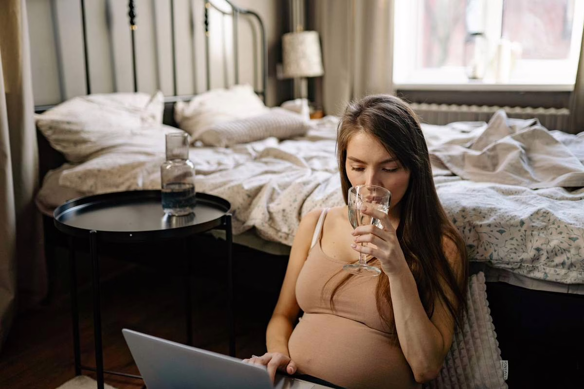 Femme enceinte au bureau buvant un verre d'eau près d'une fenêtre lors d'une journée de forte chaleur