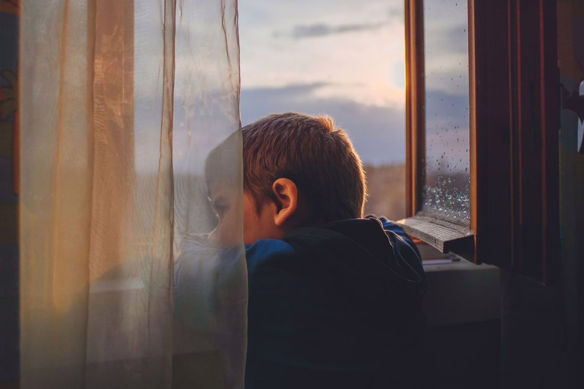Un enfant pensif assis près d'une fenêtre à la maison, illustrant le sujet de la santé mentale des plus jeunes