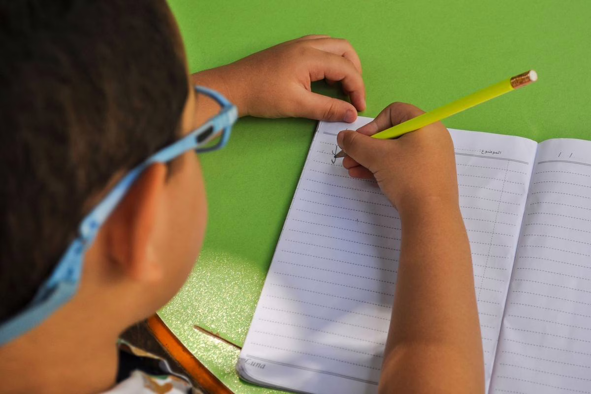Un écolier écrit des phrases complètes au crayon sur son cahier de classe, penché sur son travail