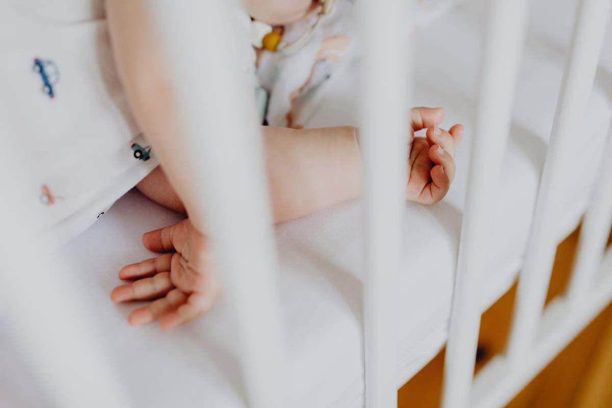 Mains et bras d'un bébé allongé dans un lit à barreaux blanc, sur un matelas ferme sans coussin ni couverture